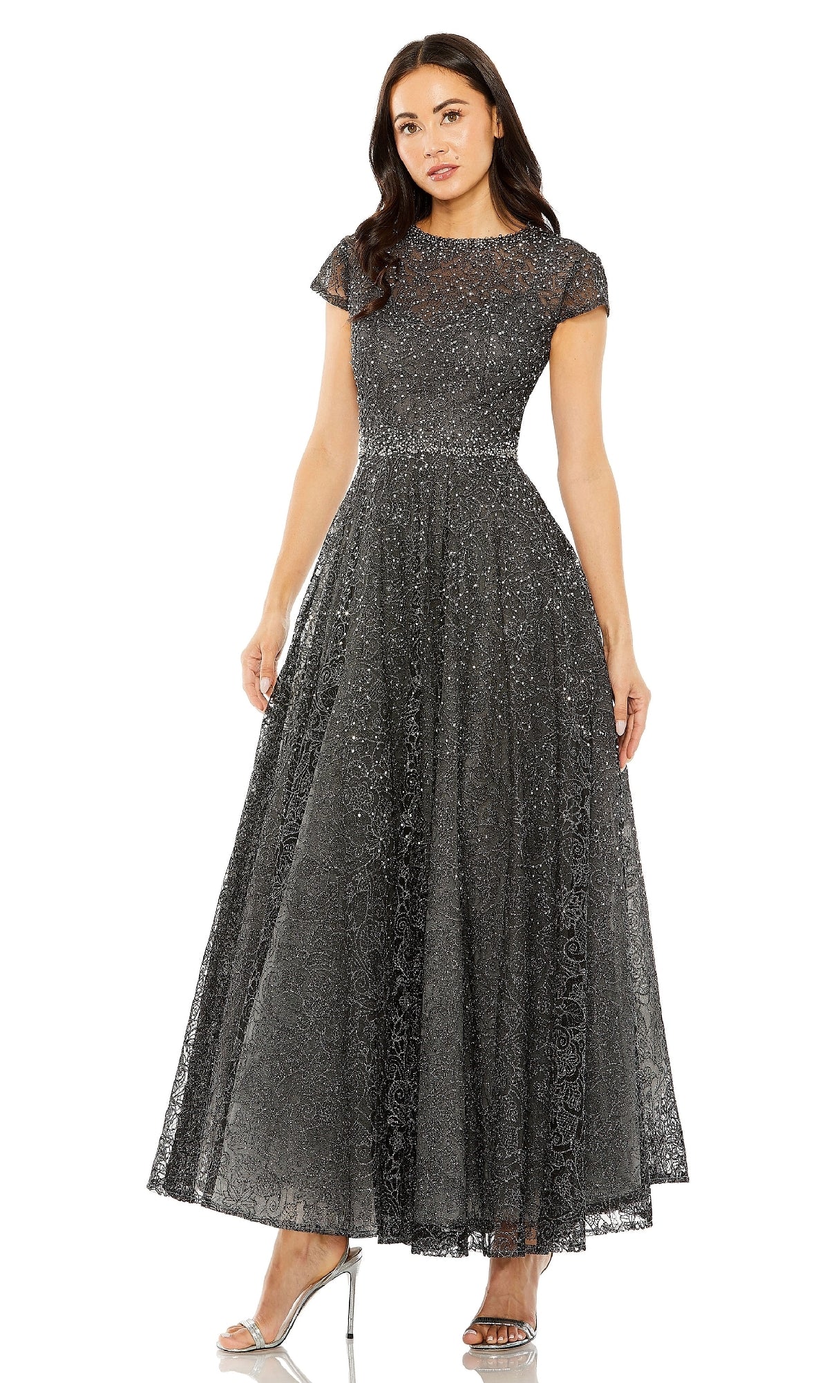 A woman models the Mac Duggal 20371 Cap Sleeve A-Line Formal Dress in dark grey lace with sparkly details and silver heels, standing against a white background.
